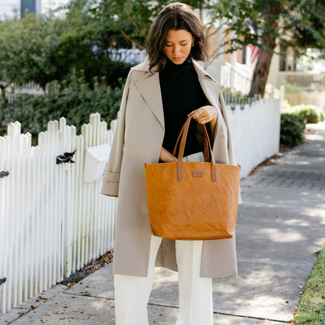 Woman holding a brown tote bag on a sidewalk with a white picket fence and greenery in the background.
