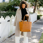 Woman holding a brown tote bag on a sidewalk with a white picket fence and greenery in the background.