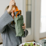 Person holding a green wine bag with a bottle over a table with food and a glass.