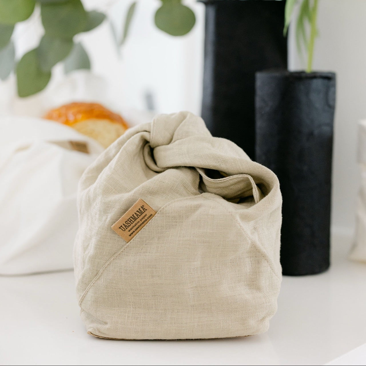 A beige linen drawstring pouch in a bright, modern interior setting with blurred green plants and black vases.