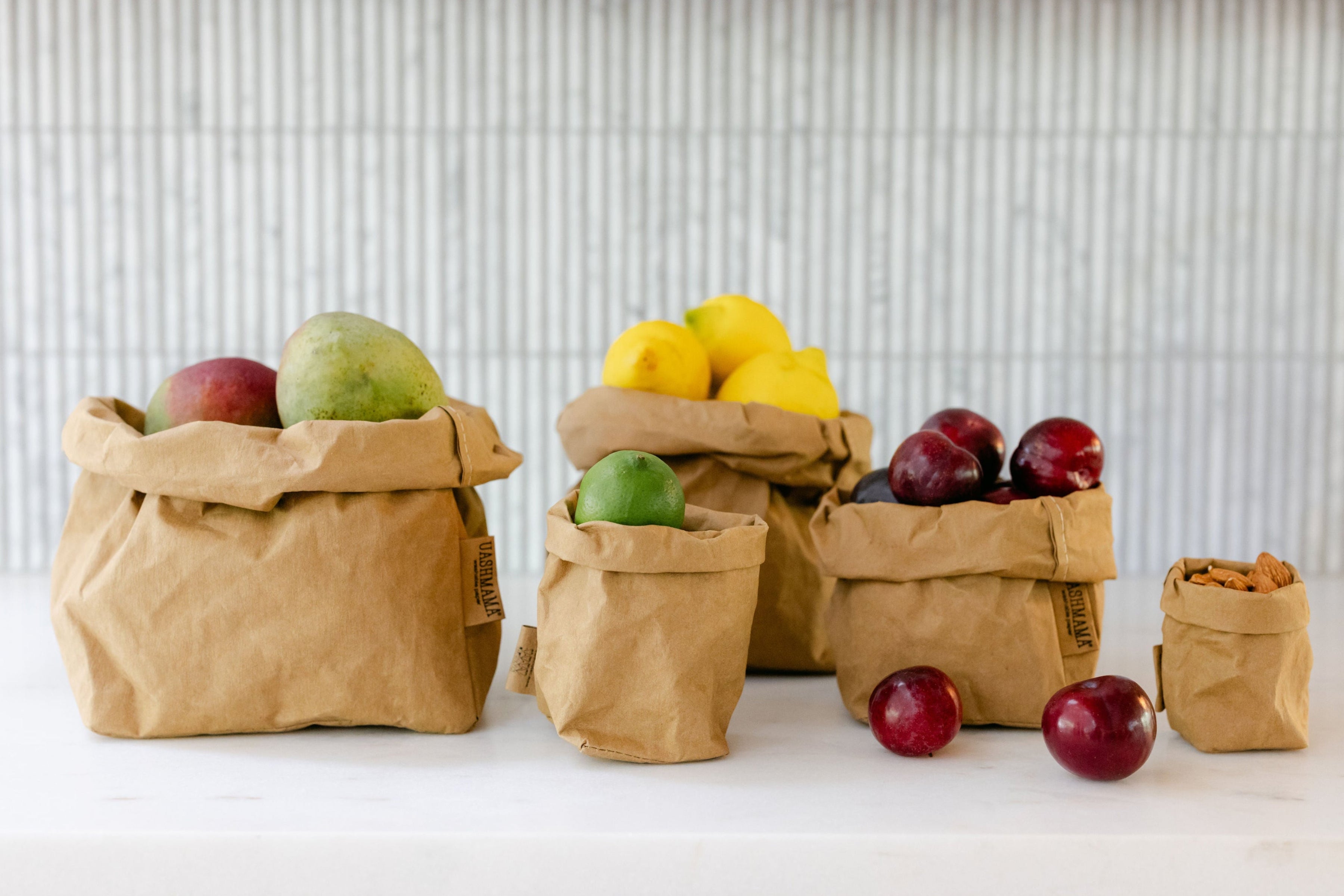 A set of brown washable paper bags filled with fresh fruits and nuts, arranged on a white surface against a light grey ribbed background.