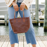Person holding a brown woven handbag on a wooden dock by water