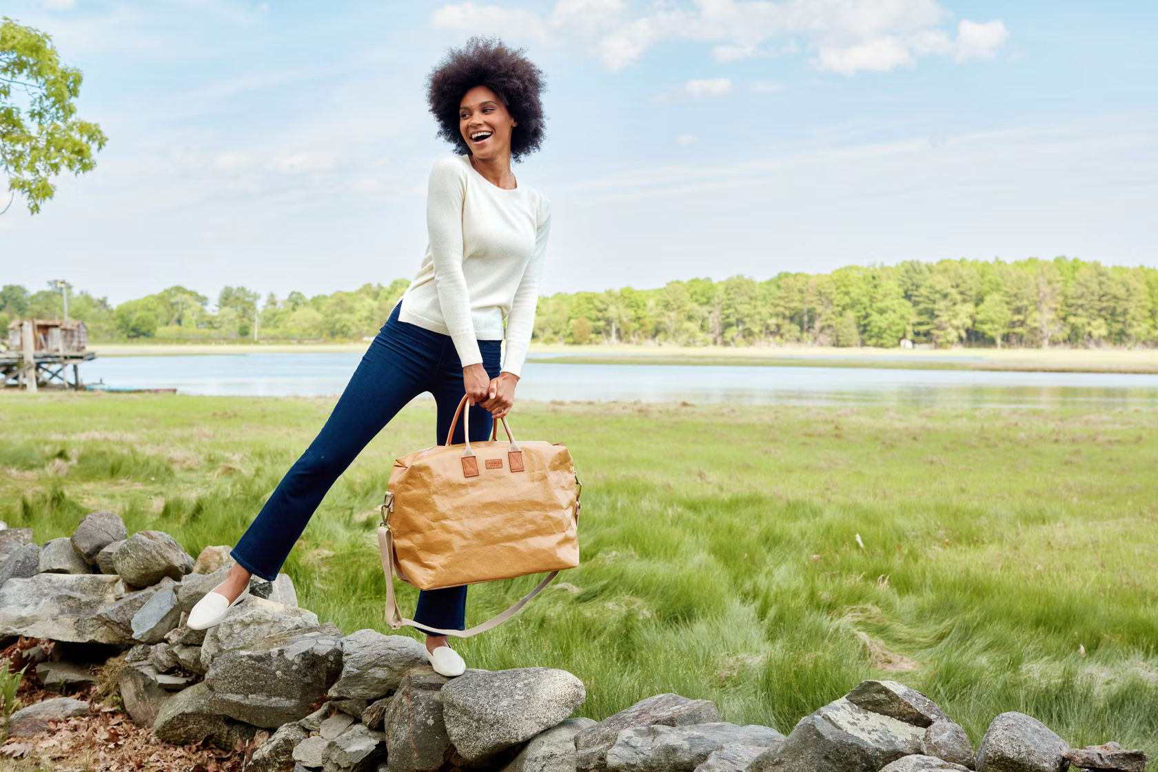 A smiling woman is shown stepping onto a rock wall. She is carrying a brown washable paper holdall.