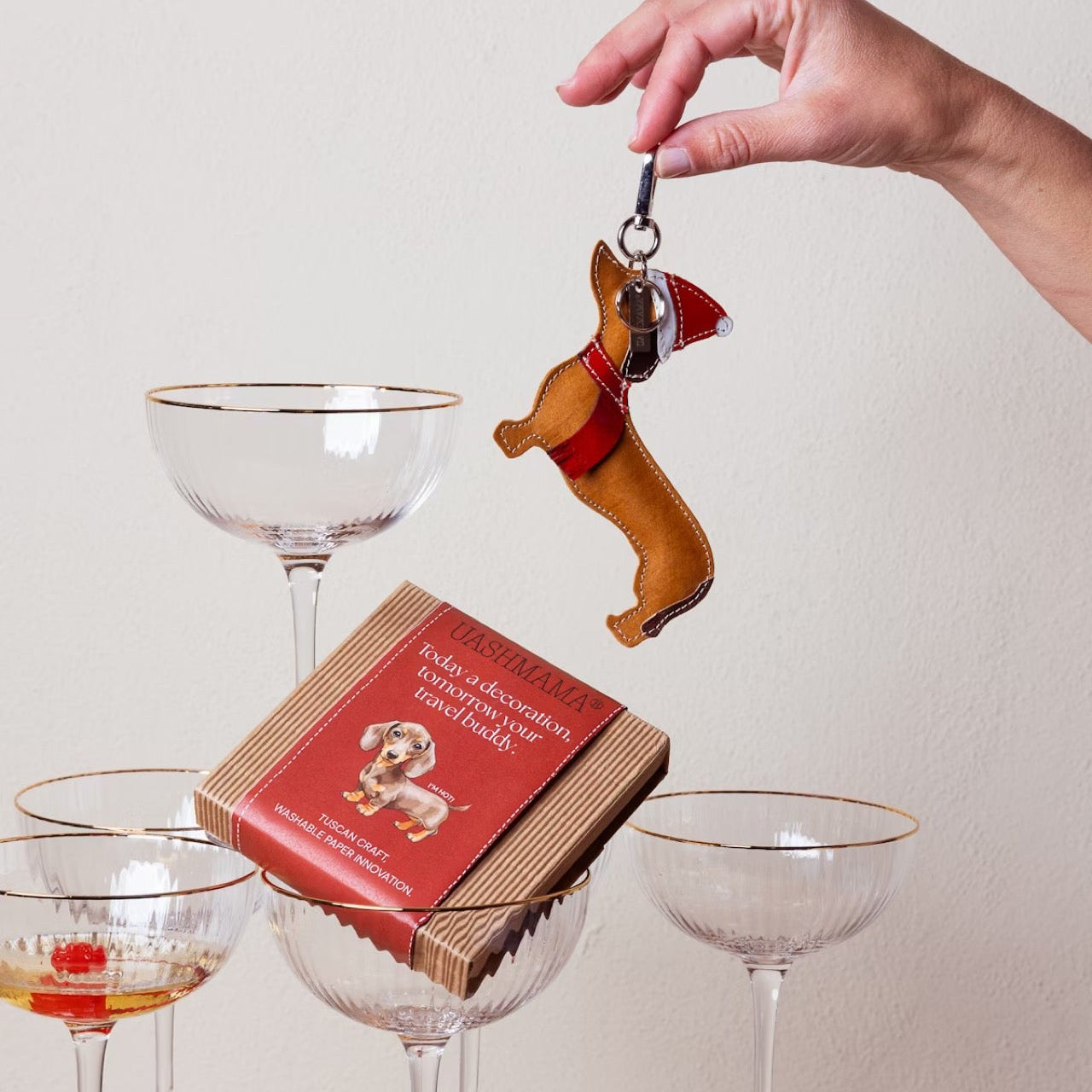 Hand holding a dog-shaped keychain above a box of chocolates with a pyramid of glasses in the background.