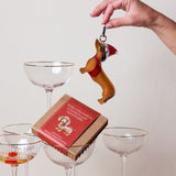 Hand holding a dog-shaped keychain above a box of chocolates with a pyramid of glasses in the background.