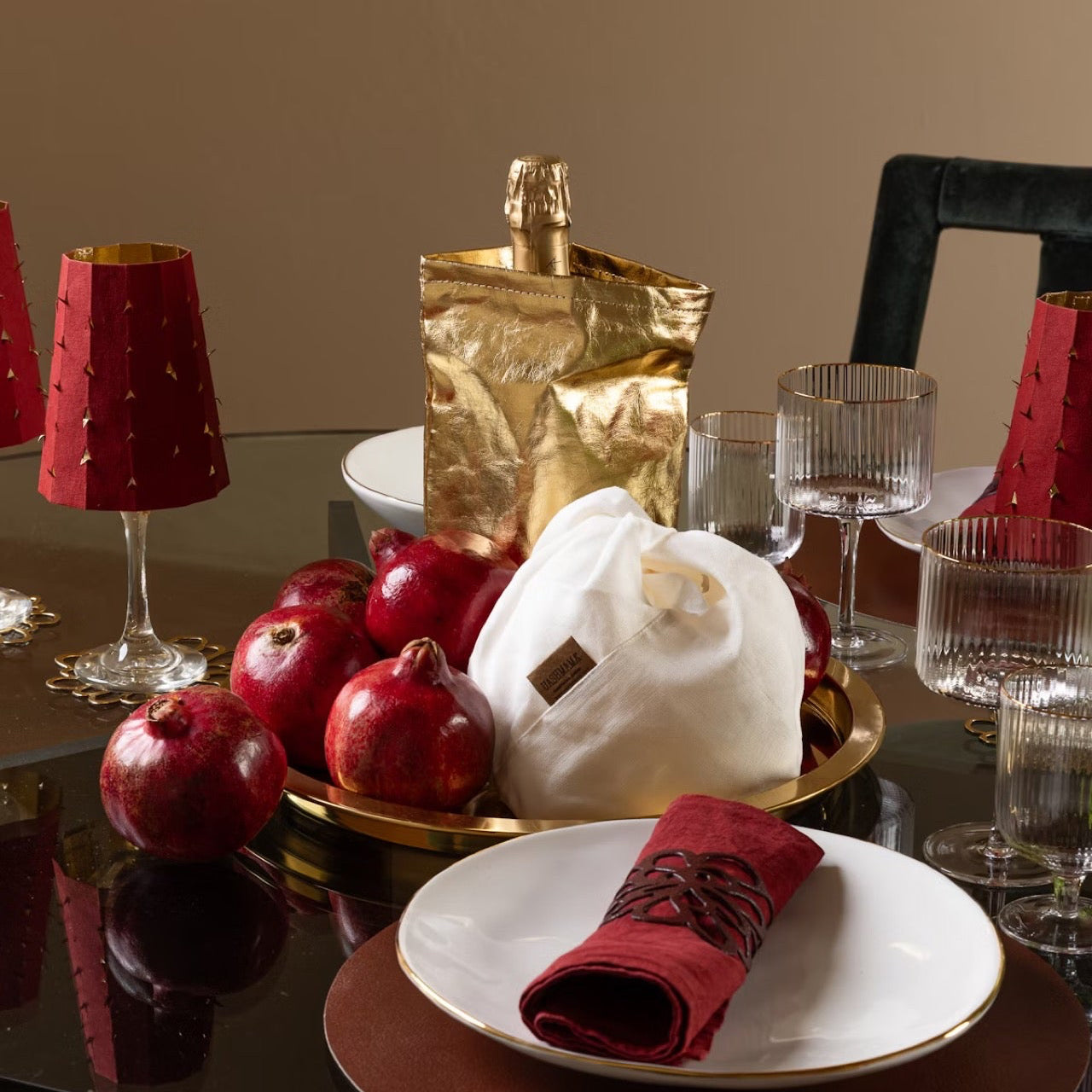 Dining table setting with red napkins, pomegranates, and a gold bag on a reflective surface.