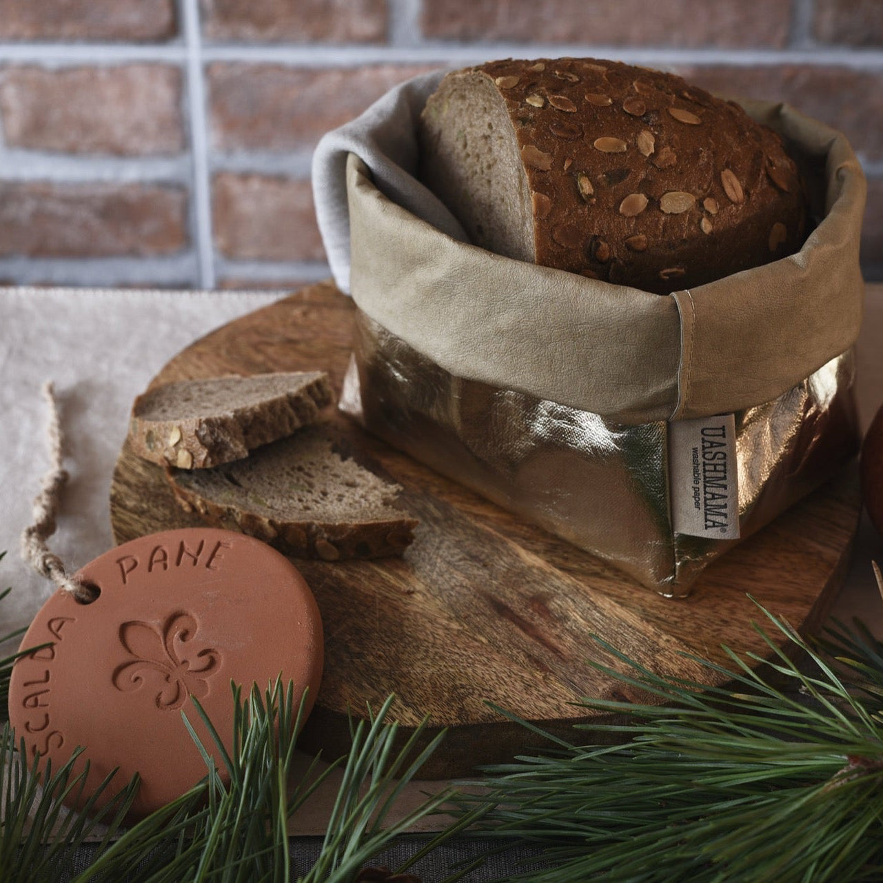 Loaf of bread in a washable paper bag on a wooden board with an apple and decorative item, against a brick wall.
