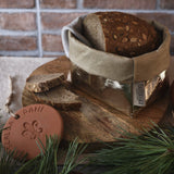 Loaf of bread in a washable paper bag on a wooden board with an apple and decorative item, against a brick wall.