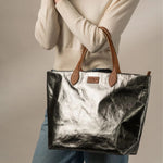 Person holding a metallic dark grey tote bag with a visible brand label against a neutral background