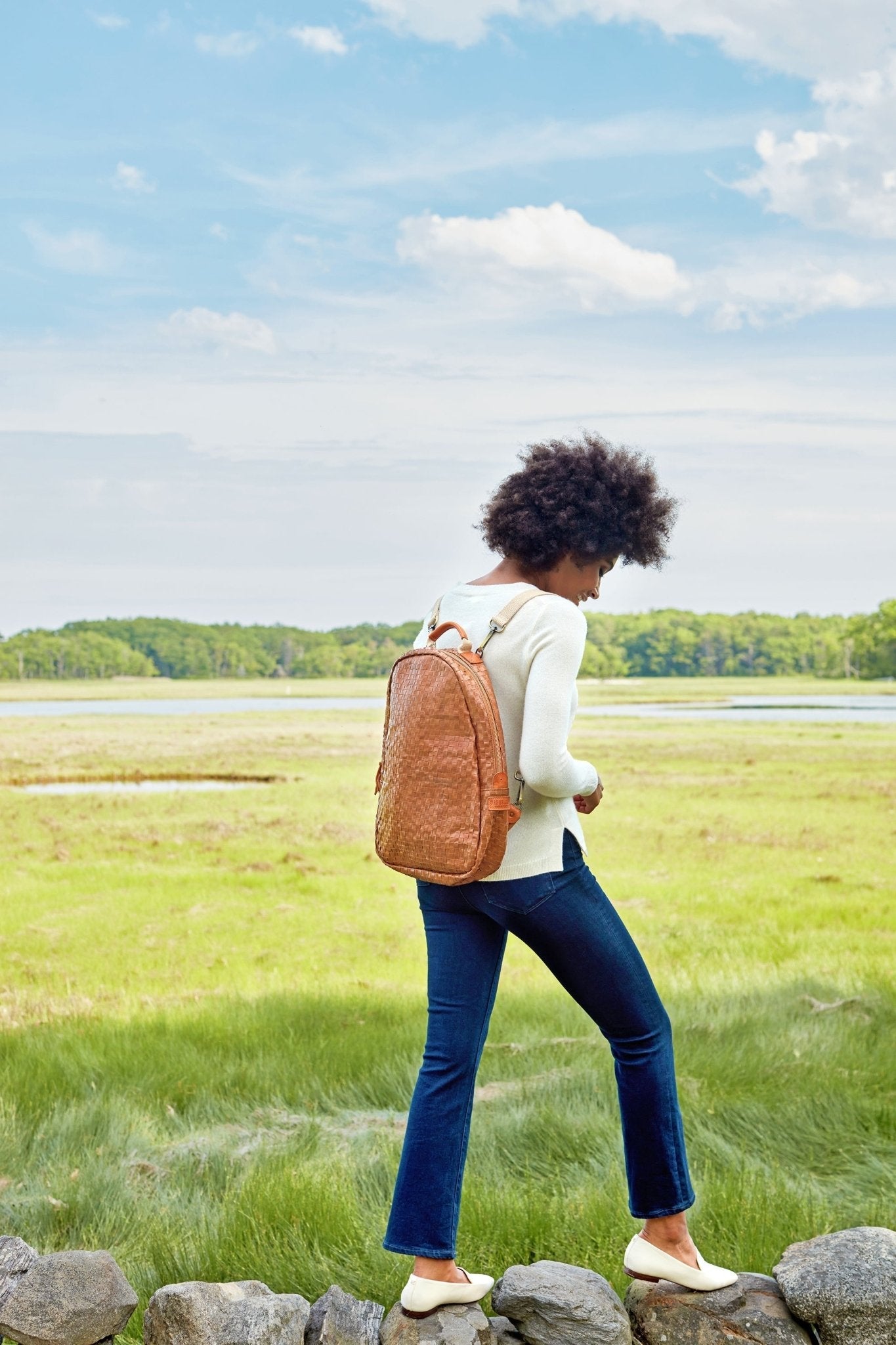 A woman is shown walking outside on top of a stone wall. She is wearing a cream jumper, blue jeans, and a tan coloured woven washable paper backpack.