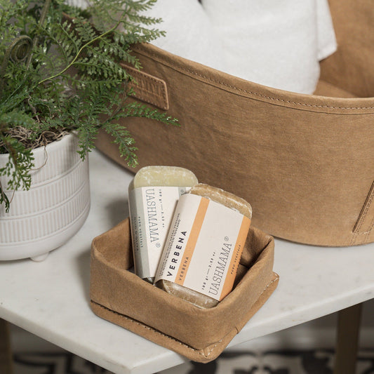 Two bars of soap (one beige, one brown) sit inside a washable paper tray. A fern sits at left and a washable paper basket is shown in the rear, containing towels.