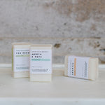 Three vertical soaps are shown on a marble shelf. The two at left are standing and the one at right is lying on its side. They are all rectangular in shape, cream in colour, and wrapped in paper labels.