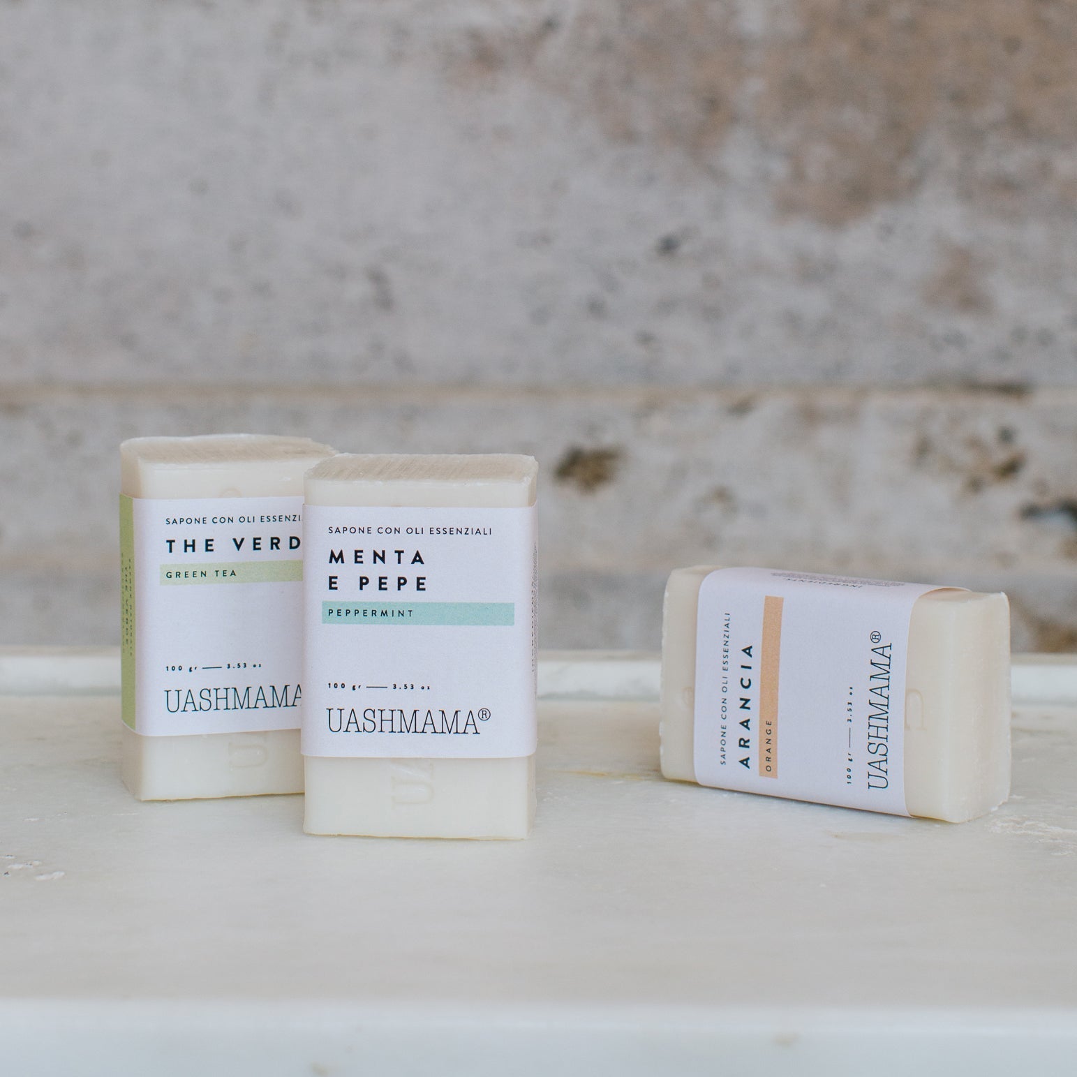 Three vertical soaps are shown on a marble shelf. The two at left are standing and the one at right is lying on its side. They are all rectangular in shape, cream in colour, and wrapped in paper labels.