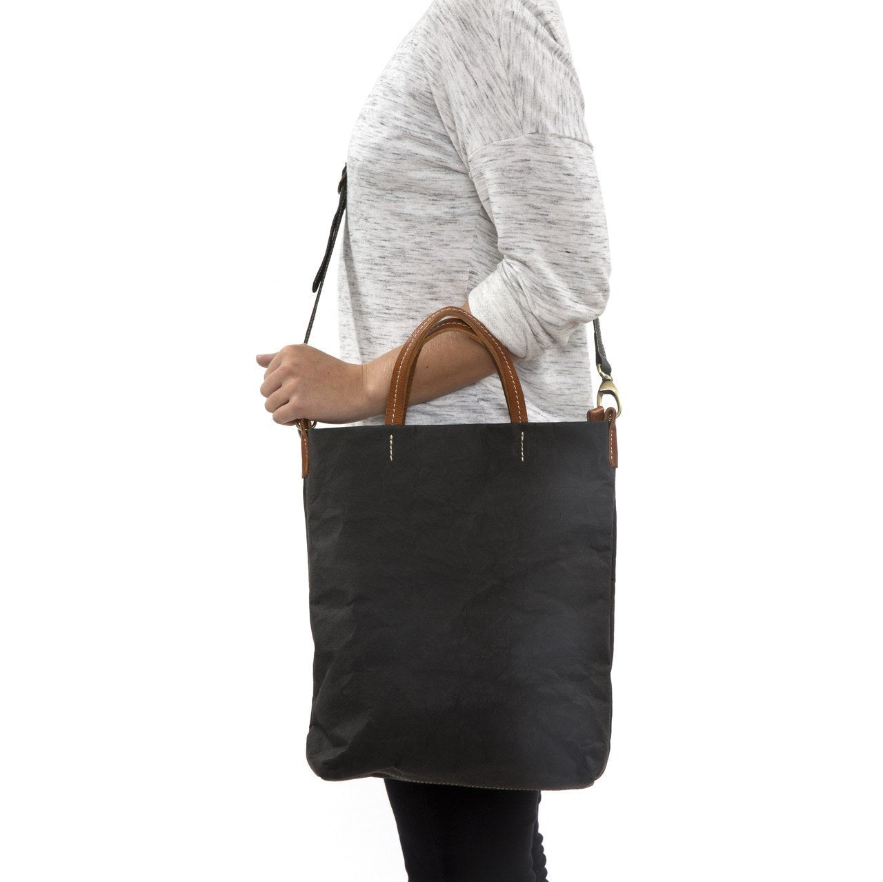 A blonde woman is shown from the side wearing casual clothing. She wears a crossbody washable paper tote bag with top handles. It is black in colour.