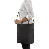 A blonde woman is shown from the side wearing casual clothing. She wears a crossbody washable paper tote bag with top handles. It is black in colour.