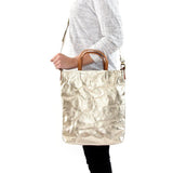 A blonde woman is shown from the side wearing casual clothing. She wears a crossbody washable paper tote bag with top handles. It is metallic gold in colour.