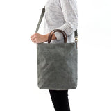 A blonde woman is shown from the side wearing casual clothing. She wears a crossbody washable paper tote bag with top handles. It is dark grey in colour.