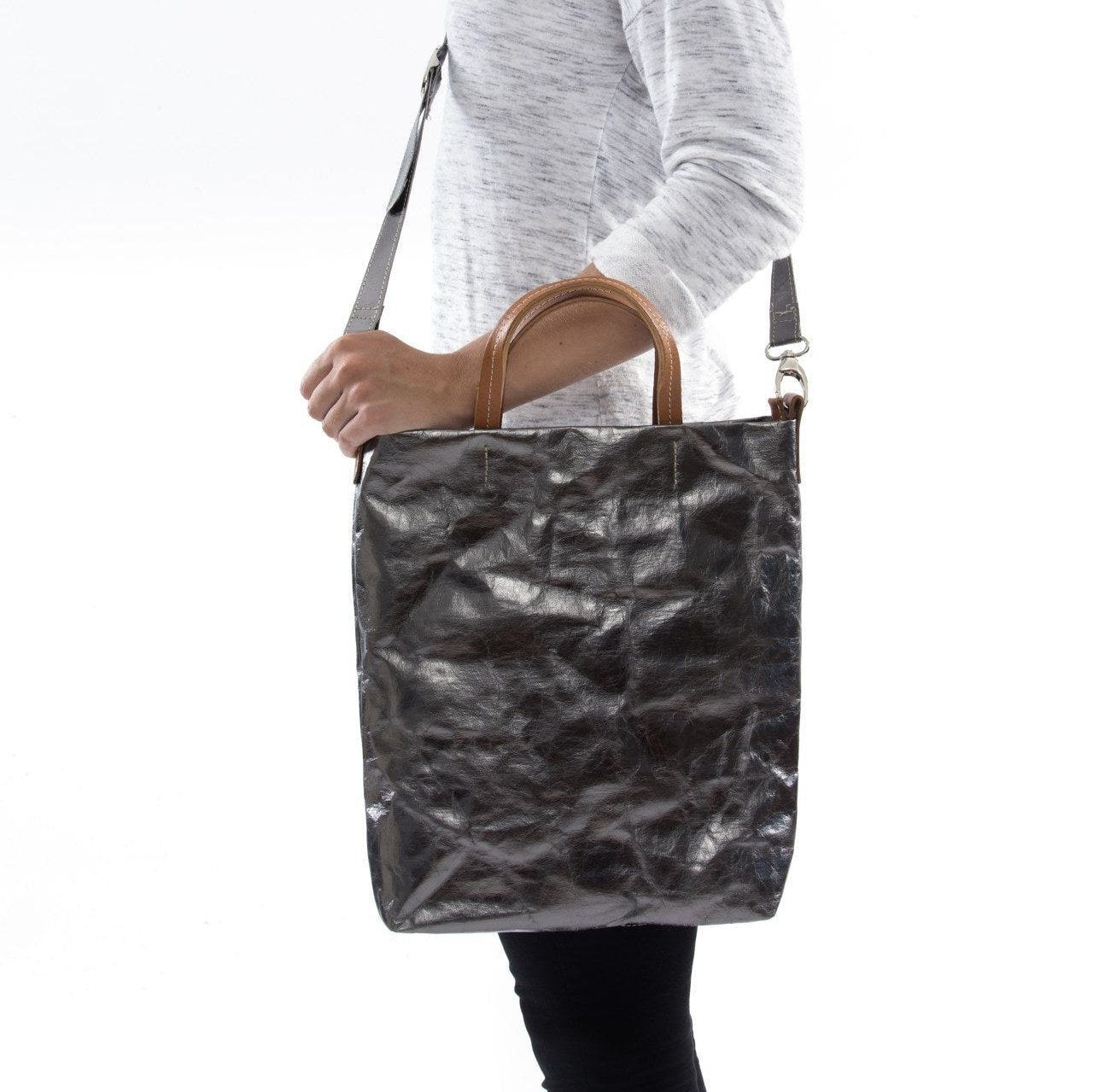 A blonde woman is shown from the side wearing casual clothing. She wears a crossbody washable paper tote bag with top handles. It is metallic pewter in colour.