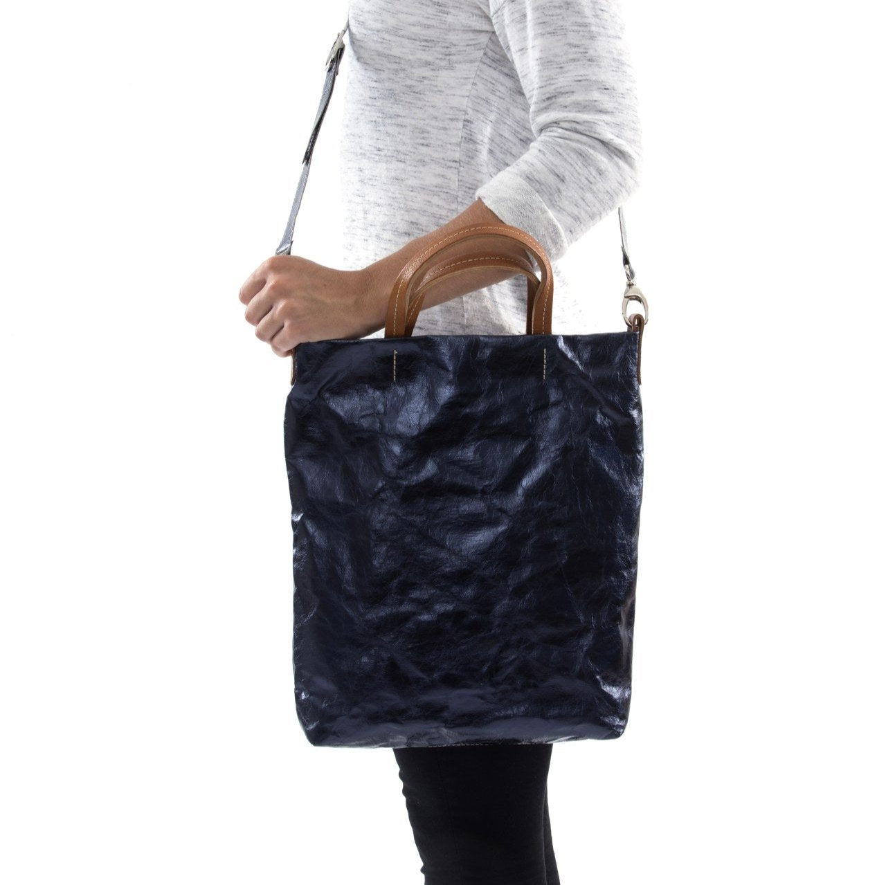 A blonde woman is shown from the side wearing casual clothing. She wears a crossbody washable paper tote bag with top handles. It is metallic navy in colour.