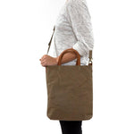 A blonde woman is shown from the side wearing casual clothing. She wears a crossbody washable paper tote bag with top handles. It is khaki in colour.