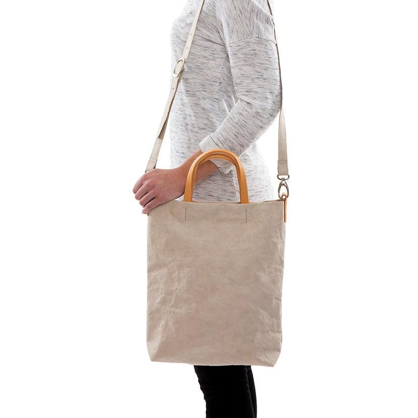 A blonde woman is shown from the side wearing casual clothing. She wears a crossbody washable paper tote bag with top handles. It is beige in colour.