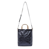 A washable paper tote is shown with a long shoulder strap pointing skywards. It has two top handles and is metallic navy in colour.