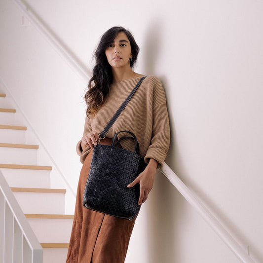 A woman stands on a white staircase wearing a beige jumper and a brown skirt. Over one shoulder she carries a black washable paper woven tote bag.