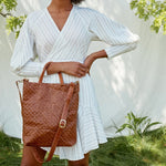 A woman is shown outdoors amidst foliage, in front of a linen backdrop. She wears a white dress and carries a brown woven washable paper tote bag over one arm.