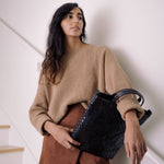 A woman stands on a white staircase wearing a beige jumper and a brown skirt. She is shown close up, with a black washable paper tote bag over one wrist.