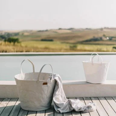 Two washable paper baskets sit outdoors on decking. The one in the front is shown in a cream colour, with a towel draped out of it. The one in the rear is smaller and shown in white.