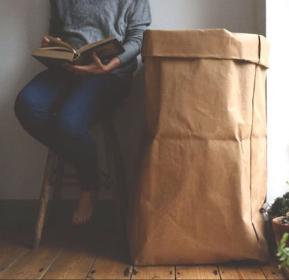 A washable paper bag is shown. The bag is rolled down at the top and features a UASHMAMA logo label on the bottom left corner. The bag pictured is the gigantic size in tan. A woman is shown sitting on a wooden stool reading a book, next to the washable paper bag. The washable paper bag comes up to the woman's elbow.