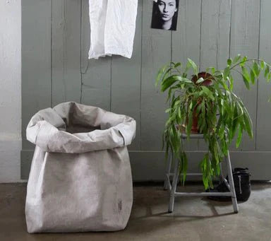 A washable paper bag is shown. The bag is rolled down at the top and features a UASHMAMA logo label on the bottom left corner. The bag pictured is the gigantic size in pale grey. Next to the bag is a metal stool with a plant on top.