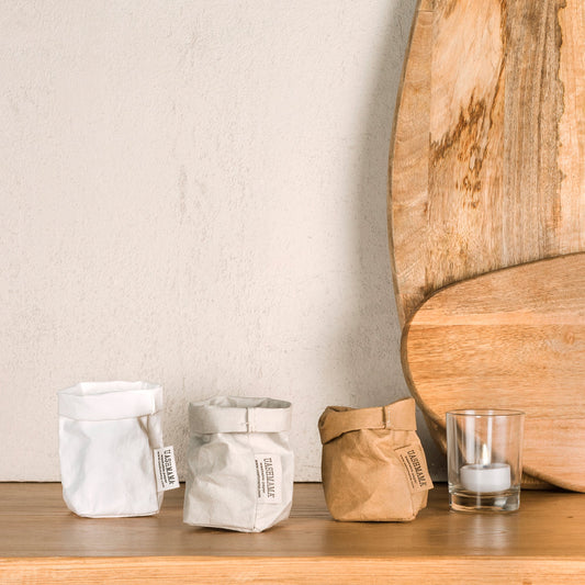 Three extra small washable paper bags are on a wooden shelf. From left to right they are white, pale grey and tan. Next to the bags is a glass votive candle. Two wooden chopping boards are leaning up against the wall in the background.