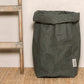 A washable paper bag is shown. The bag is rolled down at the top and features a UASHMAMA logo label on the bottom left corner. The bag pictured is the extra extra large size in dark grey. Next to the paper bag is a wooden ladder.