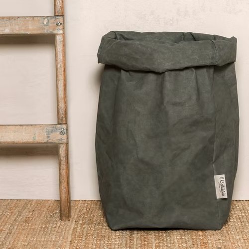 A washable paper bag is shown. The bag is rolled down at the top and features a UASHMAMA logo label on the bottom left corner. The bag pictured is the extra extra large size in dark grey. Next to the paper bag is a wooden ladder.