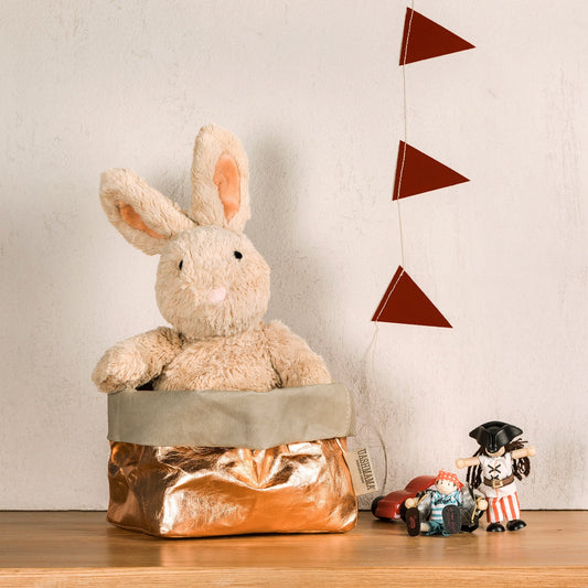 A washable paper bag is shown. The bag is rolled down at the top and features a UASHMAMA logo label on the bottom left corner. The bag pictured is the large size in rose gold. Sitting in the paper bag is a rabbit soft toy. Next to the paper bag there are two small wooden pirate toys. Hanging in the background is a string of red bunting flags hanging vertically.