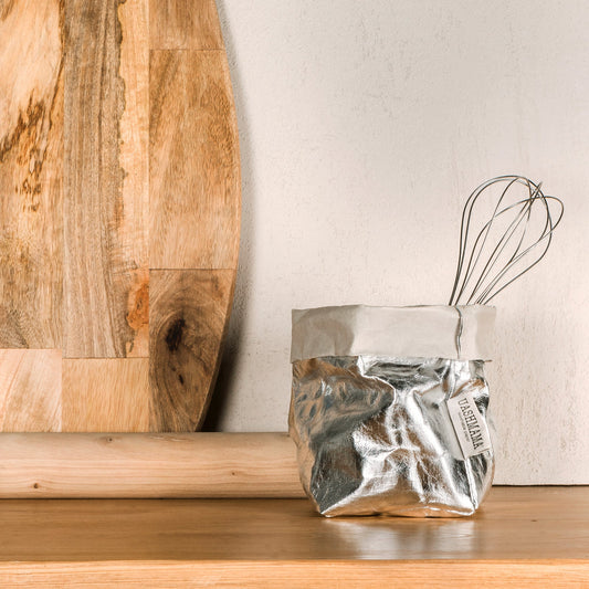 A small washable paper bag is shown in metallic silver. The bag contains a small metal whisk. Leaning up against the wall in the background is a wooden chopping board. Also shown is a wooden rolling pin.