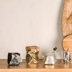 Three extra small washable paper bags are on a wooden surface. The bag on the left is dark grey metallic and empty. The bag in the middle is gold  metallic an empty. The bag on the right is platinum metallic an contains a teaspoon. To the right of the washable paper bags are two glass espresso mugs. Two wooden chopping boards are shown leaning up against the wall.