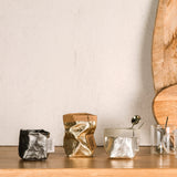 Three extra small washable paper bags are on a wooden surface. The bag on the left is dark grey metallic and empty. The bag in the middle is gold  metallic an empty. The bag on the right is platinum metallic an contains a teaspoon. To the right of the washable paper bags are two glass espresso mugs. Two wooden chopping boards are shown leaning up against the wall.