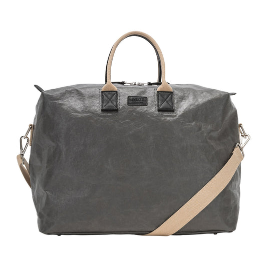A washable paper holdall with two short carry handles and one long fabric strap. The holdall closes with a zip which has leather zip pulls. The holdall has leather details attaching the straps to the bag and a small UASHMAMA leather logo label. The bag shown is dark grey with wheat coloured handles and strap. The holdall is extra large in size.