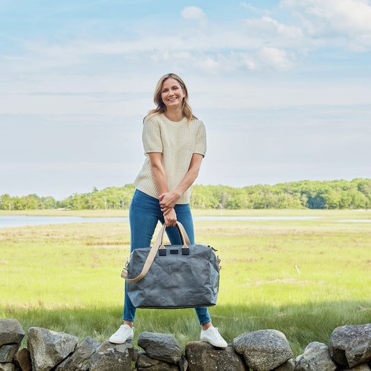 A woman is shown standing on a stone wall with a grass field in the background. She is holding a dark grey washable paper holdall with wheat coloured handles and shoulder strap.