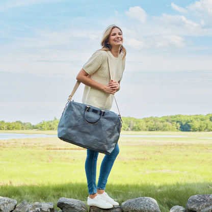 A woman is shown standing on a stone wall with a grass field in the background. On her shoulder she is carrying a dark grey washable paper holdall with wheat coloured handles and shoulder strap.