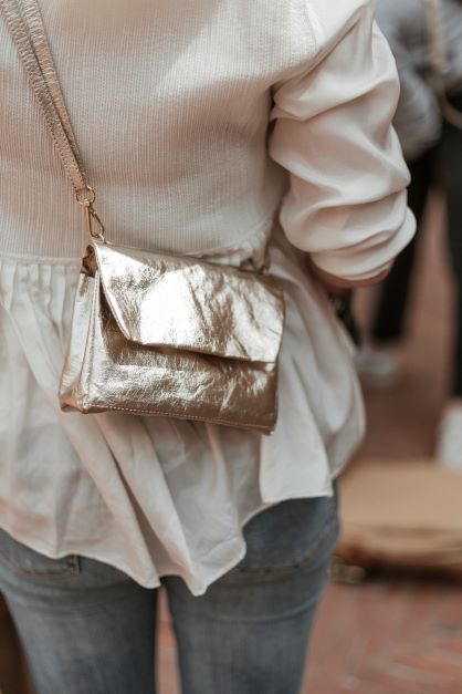 The torso of a woman is shown. She is wearing jeans and a cream sweater, and she is facing away from the camera. Across her body she is wearing a small washable paper handbag with a front flap closure. The bag is metallic platinum in colour.