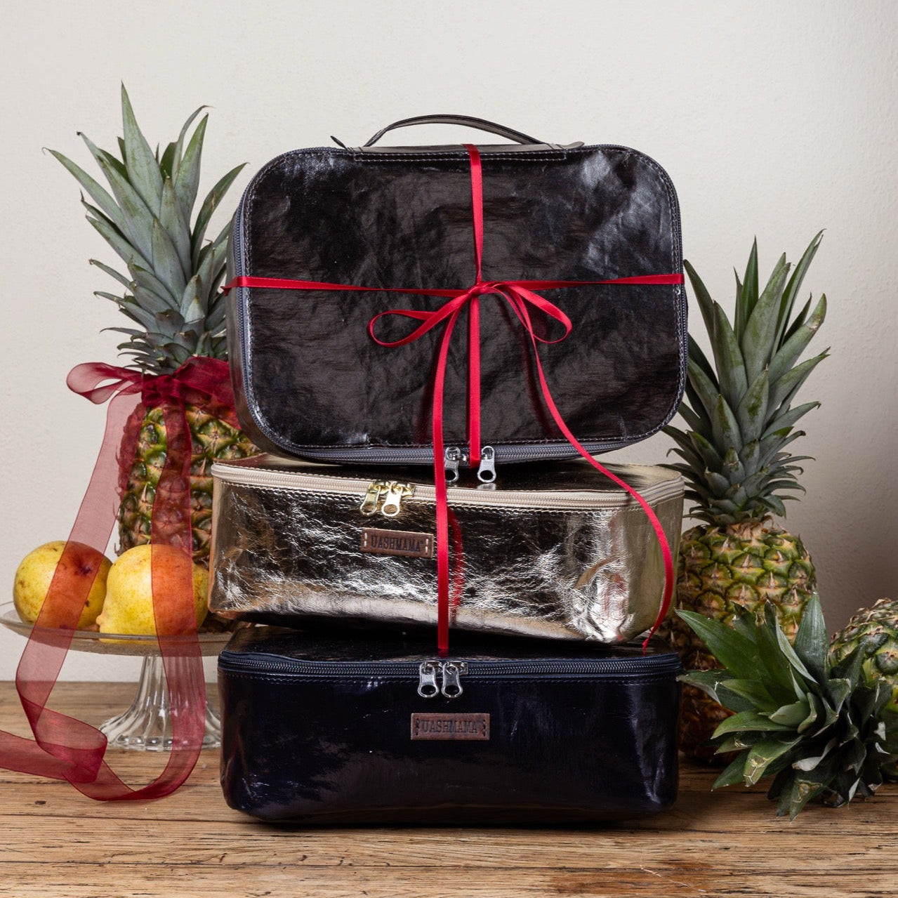 Set of black and silver suitcases with red ribbons on a wooden surface with decorative fruits and plants.
