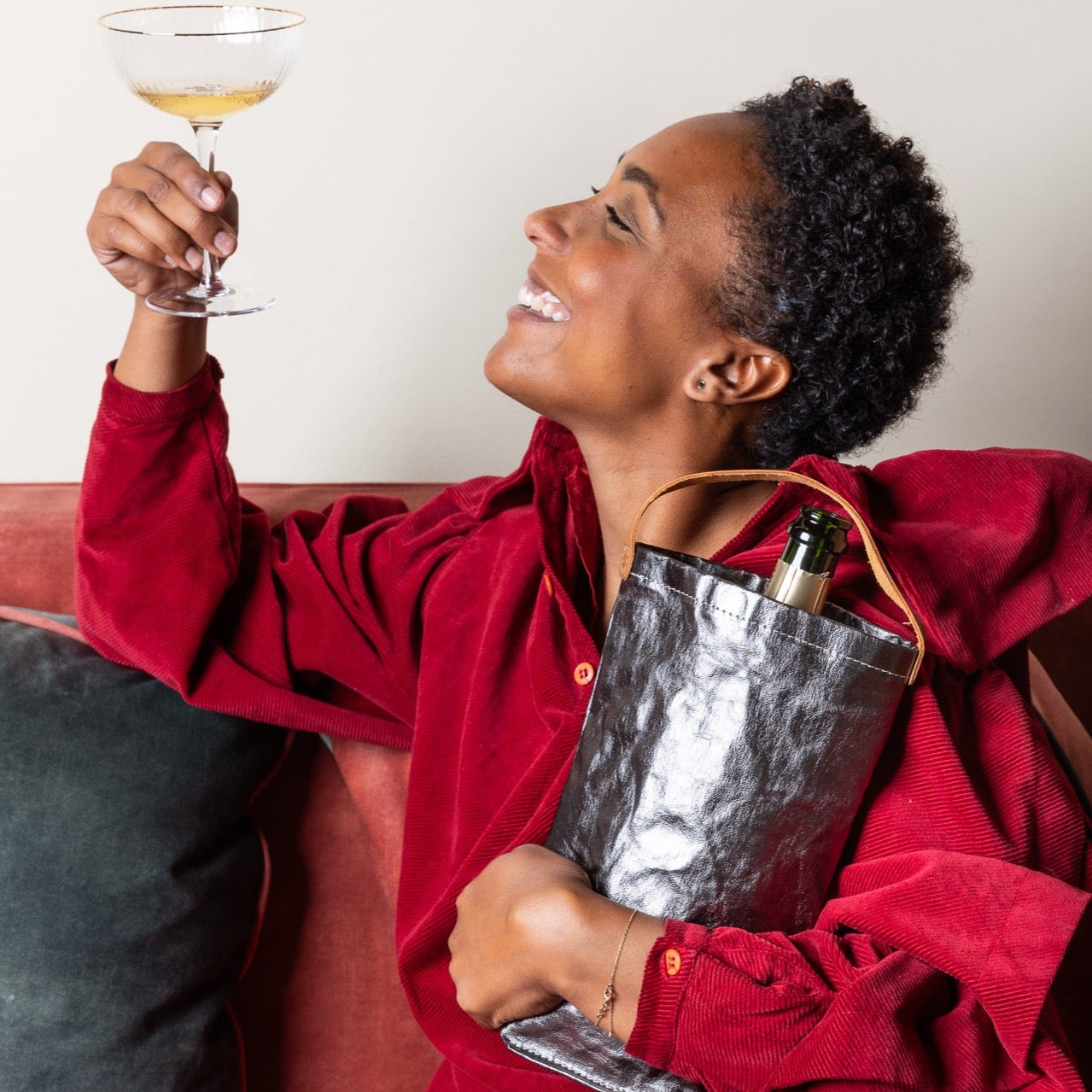 Person in a red outfit holding a cocktail and a metallic bag on a couch.