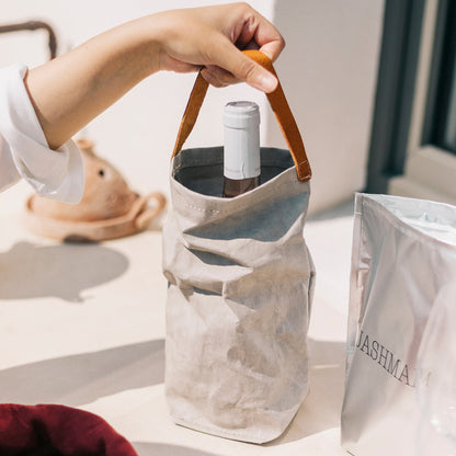 A woman's hand is shown holding a pale grey washable paper wine bag by its tan leather handle. Next to the wine bag on the table is a silver UASHMAMA branded wine cooler.