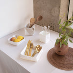 A series of washable paper trays and holders are shown on a white table top. The white tray at left contains fruit and the white tray at right holds gold cutlery.