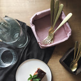 A washable paper tray is shown in pink, containing gold cutlery. At right sits a smaller brown washable paper tray containing gold teaspoons. A cake sits on a plate at left.