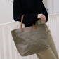 A woman is shown leaning against a white railing. She is wearing a large washable paper tote bag. It has two long tan leather handles. There is a small UASHMAMA logo leather label on the front of the bag in between the handles. The bag shown is in an olive colour.