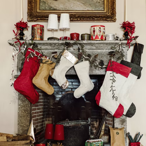A grey marble fireplace is shown with Christmas decorations and tins on the mantelpiece. Hanging above the fire are three small and three large washable paper stockings in various colours. 
